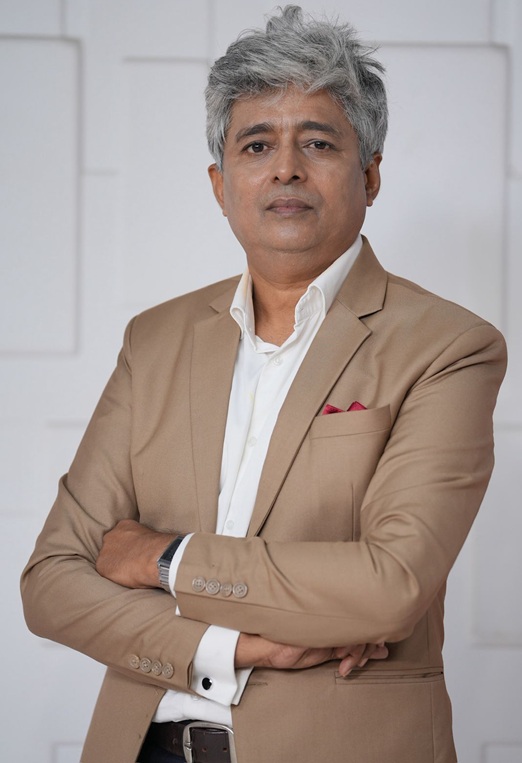 Professional portrait of a middle-aged man with gray hair, wearing a tan suit and white shirt, arms crossed against a light, neutral background.