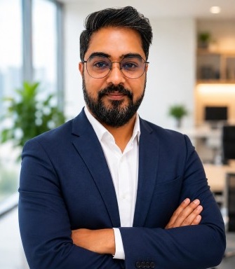 Professional man with dark hair and beard in a navy suit, arms crossed in a bright office.