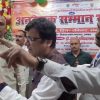 Group of men at a formal event, two in the foreground pointing and discussing as others listen, with a Hindi banner in the background.