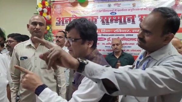 Group of men at a formal event, two in the foreground pointing and discussing as others listen, with a Hindi banner in the background.