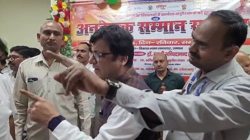 Group of men at a formal event, two in the foreground pointing and discussing as others listen, with a Hindi banner in the background.