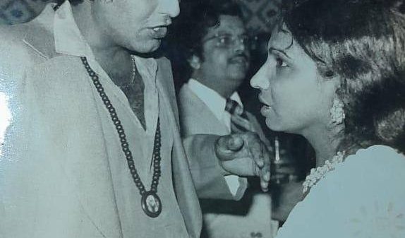 Man in a light suit speaking to a woman in a white dress at a social gathering; vintage setting with a guest in the background.