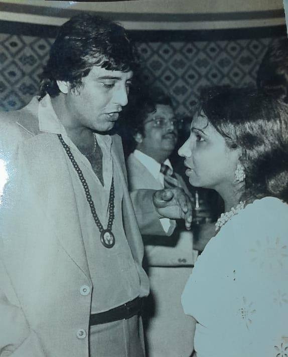 Man in a light suit speaking to a woman in a white dress at a social gathering; vintage setting with a guest in the background.