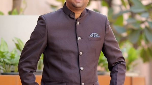 Smiling man in a dark brown mandarin-collar suit posing outdoors with plants in the background.