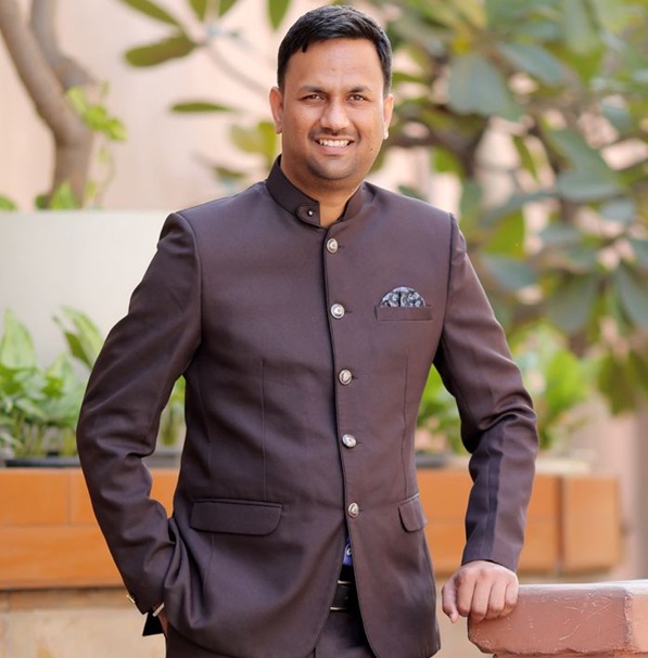 Smiling man in a dark brown mandarin-collar suit posing outdoors with plants in the background.