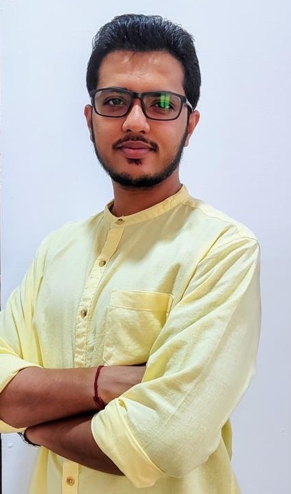 Man with short dark hair and glasses wearing a pale yellow kurta, arms crossed, looking at the camera.