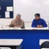 Two men sit at a panel desk at the front of a classroom, one speaking into a microphone while the other listens.