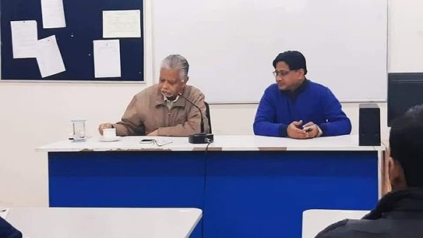 Two men sit at a panel desk at the front of a classroom, one speaking into a microphone while the other listens.