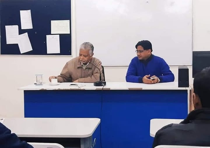 Two men sit at a panel desk at the front of a classroom, one speaking into a microphone while the other listens.