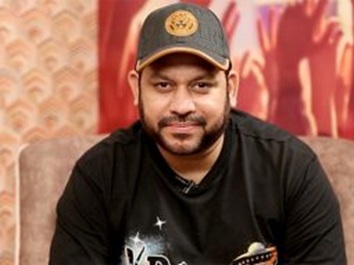 Man with a beard wearing a black T-shirt and gray cap, seated on a sofa with a patterned backdrop.