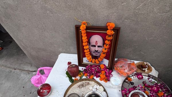 Framed portrait on a small roadside altar with orange marigold garlands, surrounded by fruit, flowers, and ritual bowls on a white cloth.
