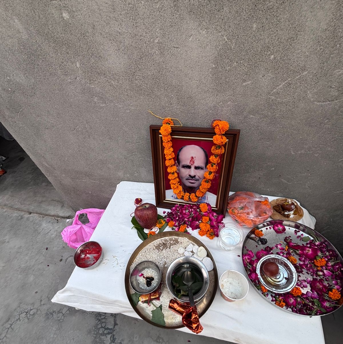 Framed portrait on a small roadside altar with orange marigold garlands, surrounded by fruit, flowers, and ritual bowls on a white cloth.