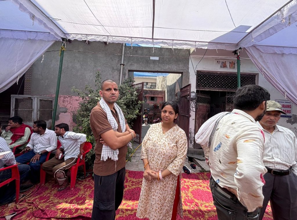 Man with a towel over his shoulder and arms crossed stands beside a woman in a floral kurta under a white canopy at an outdoor gathering.
