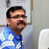Man with glasses and mustache wearing a blue striped polo shirt, seated indoors in a neutral room setting.