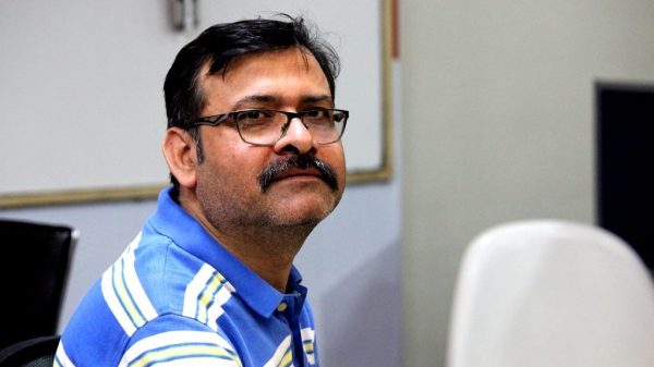 Man with glasses and mustache wearing a blue striped polo shirt, seated indoors in a neutral room setting.
