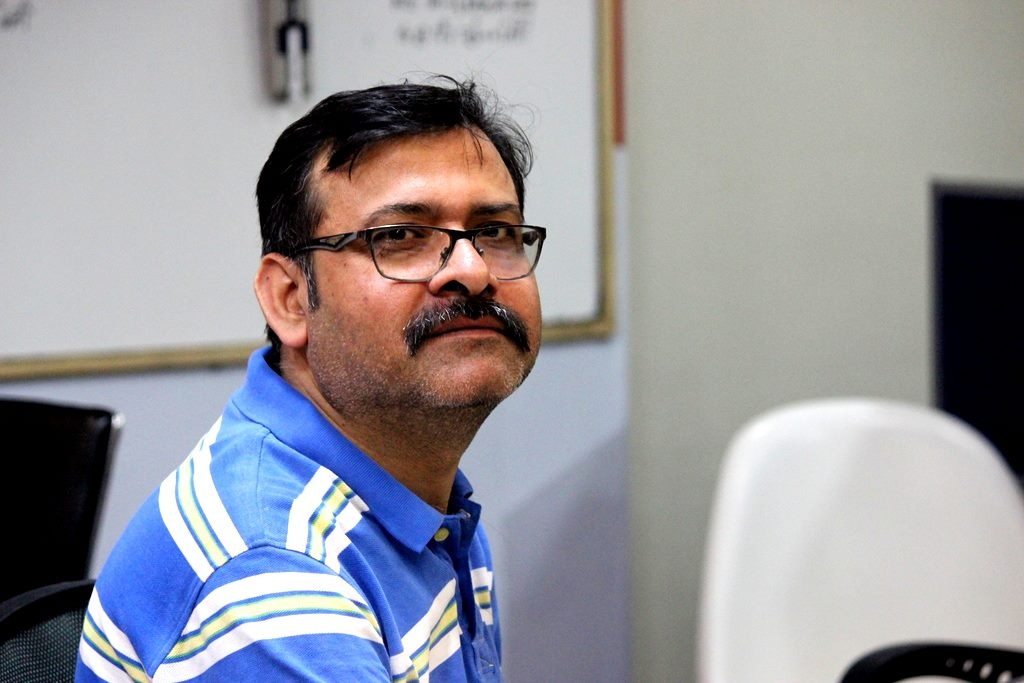 Man with glasses and mustache wearing a blue striped polo shirt, seated indoors in a neutral room setting.