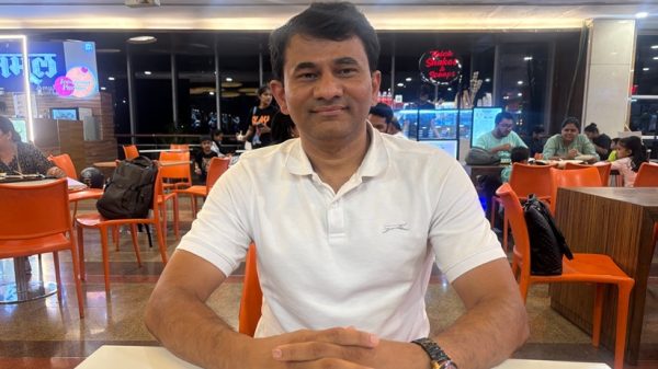 Man in a white polo sits at a table in a busy cafe, smiling at the camera.