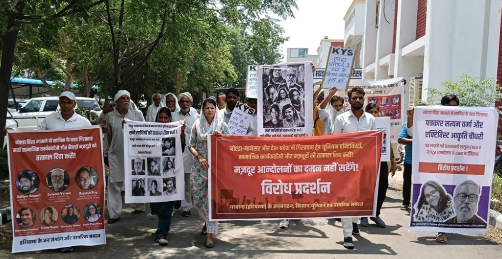 satyam_haryana | No. 1 Indian Media News Portal A group of protesters marches down a sunny street, carrying large red banners and multiple posters with Hindi text and photos. They appear focused and organized.