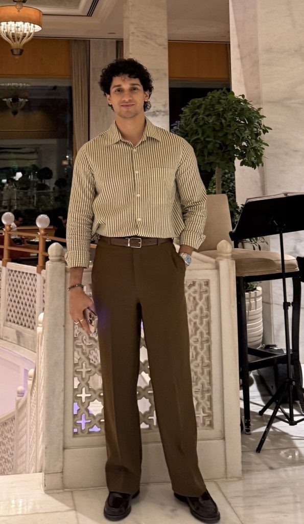 Man posing in a striped beige shirt and brown trousers indoors, hands in pockets, in an elegant venue.",