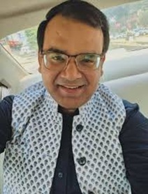 Smiling man with glasses wearing a patterned white vest, seated in a car.