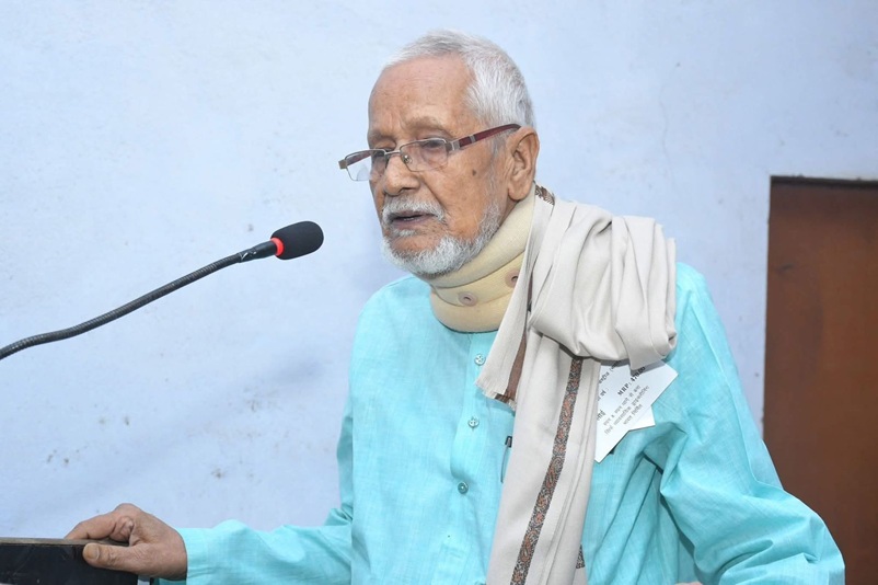 Older man with white hair and beard speaking into a microphone at a podium, wearing a turquoise kurta and scarf.