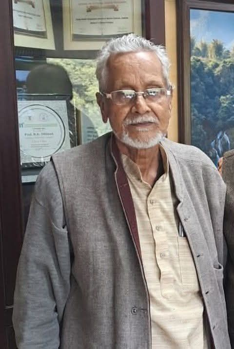 Older man with grey hair and beard wearing glasses, beige shirt and grey blazer, standing indoors with framed certificates behind him.