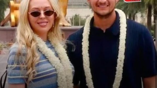 Two smiling young adults wearing flower garlands pose together outdoors, with a Hindi news headline overlaid at the bottom.