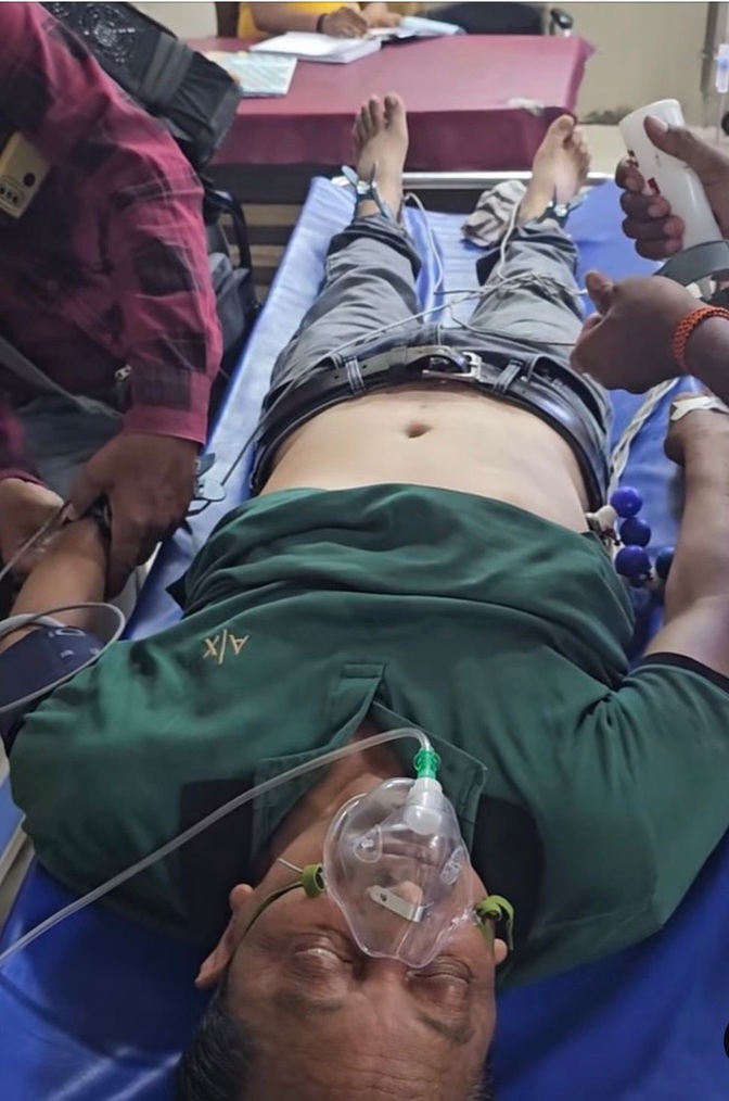 Patient lying on a hospital bed wearing a green shirt with an oxygen mask and medical tubes, surrounded by staff attending to him.