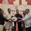 Stage with a banner reading 'गाजीपुर प्रेस क्लब' as a group of men awards a circular plaque to a man in black, with others watching and floral arrangements in front.