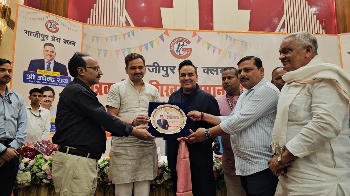 Stage with a banner reading 'गाजीपुर प्रेस क्लब' as a group of men awards a circular plaque to a man in black, with others watching and floral arrangements in front.