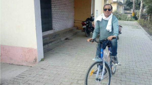 Man wearing sunglasses rides a blue bicycle along a brick-paved path beside a residential building, with a towel draped over his shoulders.
