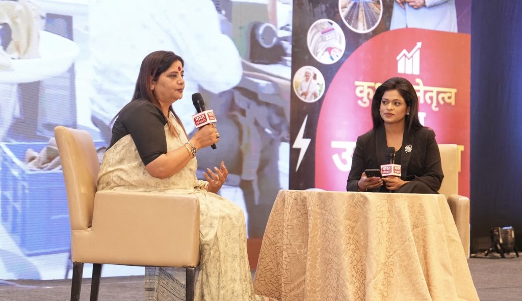 49788c0f-bab9-48a2-b65b-204b12b95ba2 | No. 1 Indian Media News Portal Two women seated on a stage in a panel discussion; the left woman speaks into a microphone while the right woman looks on, backdrop shows event graphics.