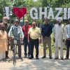 Group of men posing in front of a large 'I heart Ghazipur' sign in a park; one man stands with a bicycle in the center left.