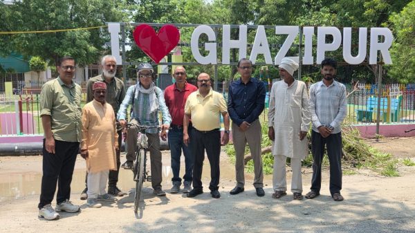 Group of men posing in front of a large 'I heart Ghazipur' sign in a park; one man stands with a bicycle in the center left.
