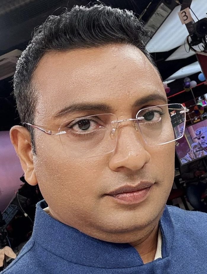 Close-up portrait of a man wearing clear rimless glasses and a blue collared shirt, looking slightly to the right.