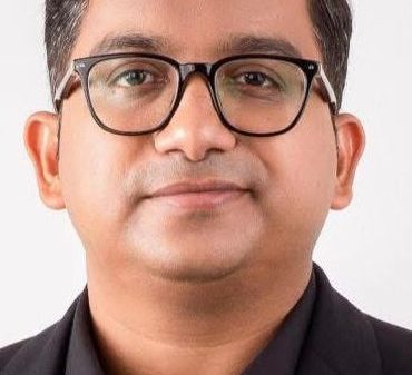Portrait of a man wearing black-rimmed glasses and a black blazer against a light background, neutral facial expression.