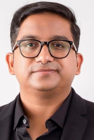Portrait of a man wearing black-rimmed glasses and a black blazer against a light background, neutral facial expression.