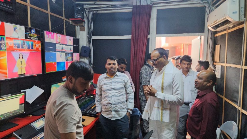 aaj ki khabar | No. 1 Indian Media News Portal A group of men stands around a broadcast control console in a studio, with large multi-screen displays showing a news anchor and color panels in the background.
