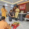 A group of people performing a Hindu household ritual on the office floor with a woman in a sari offering a plate of flowers and lamps; others stand nearby.