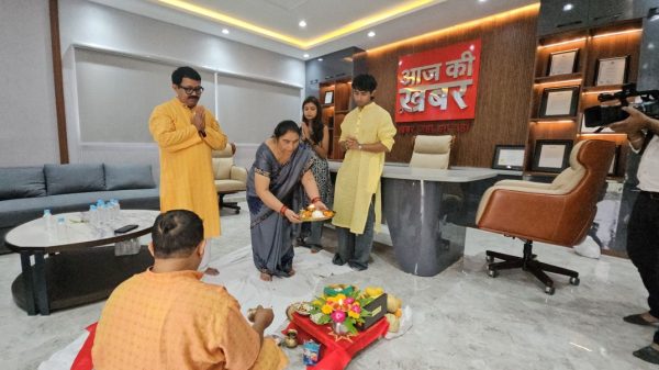 A group of people performing a Hindu household ritual on the office floor with a woman in a sari offering a plate of flowers and lamps; others stand nearby.