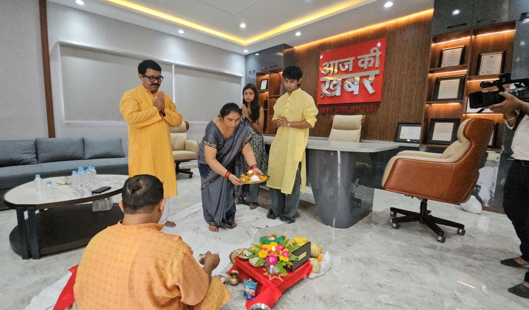 A group of people performing a Hindu household ritual on the office floor with a woman in a sari offering a plate of flowers and lamps; others stand nearby.