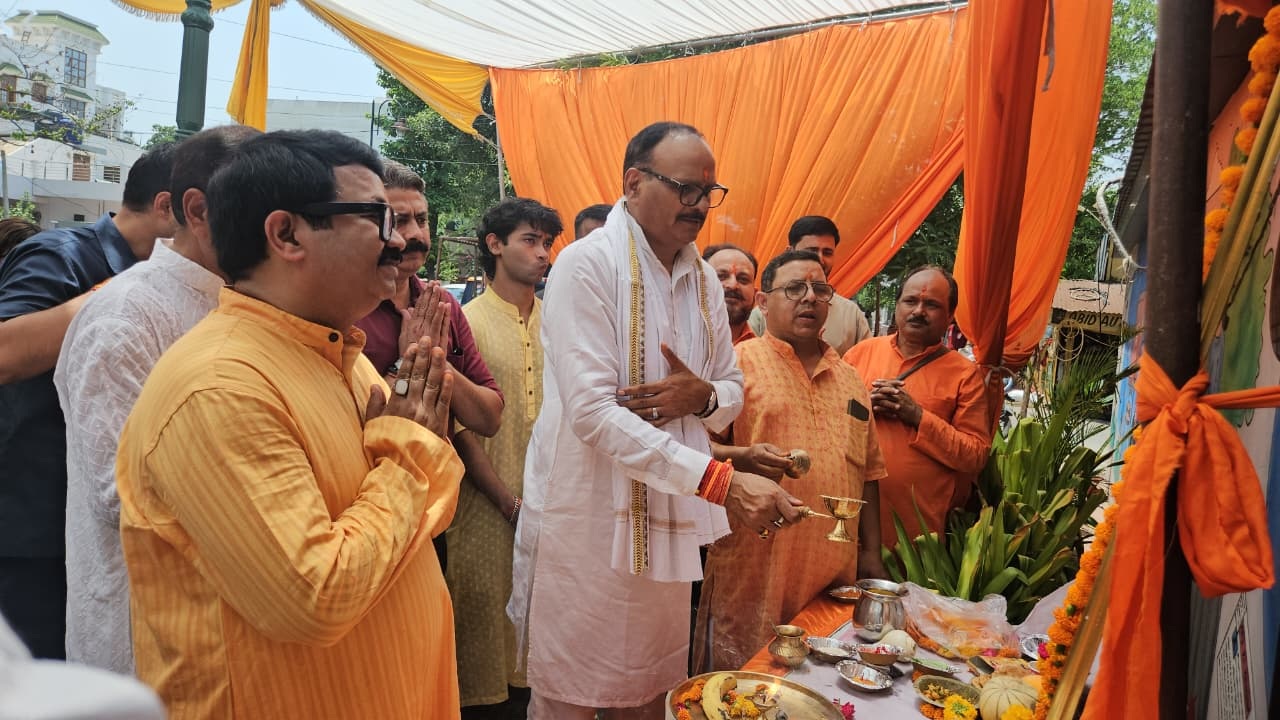 Men perform a Hindu ritual with offerings on a table under orange drapes at a ceremonial gathering.