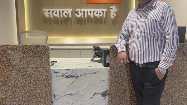 Man in a striped shirt stands at a marble reception desk in an office lobby with a backlit IBM24 sign behind him.