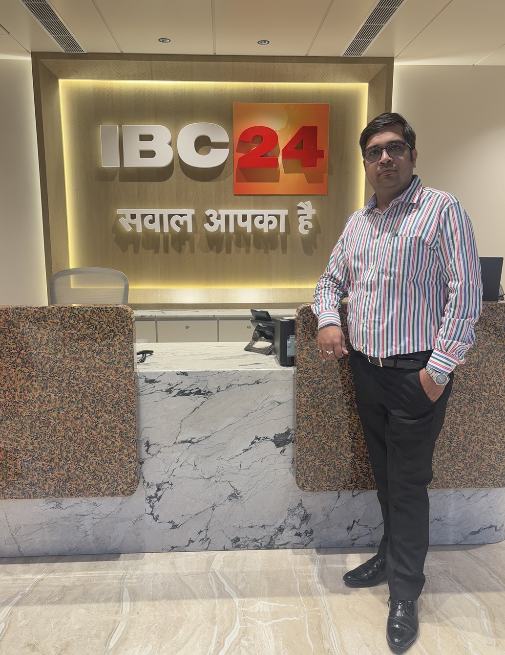 Man in a striped shirt stands at a marble reception desk in an office lobby with a backlit IBM24 sign behind him.
