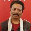 Man with dark hair and a mustache wearing a red shirt and white scarf against a red backdrop.