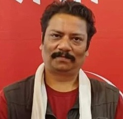 Man with dark hair and a mustache wearing a red shirt and white scarf against a red backdrop.