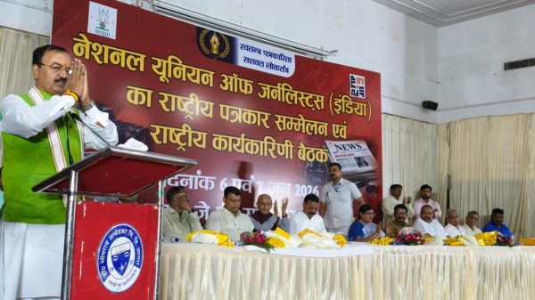 Man in a green vest speaks at a podium during a press conference or formal event, with a large red banner in Hindi behind him and a row of officials seated on stage.