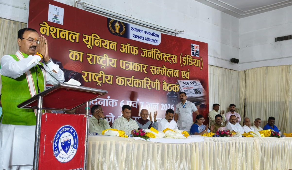 Man in a green vest speaks at a podium during a press conference or formal event, with a large red banner in Hindi behind him and a row of officials seated on stage.