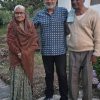 Three older adults stand together outdoors in a garden, posing for the photo in front of a house.