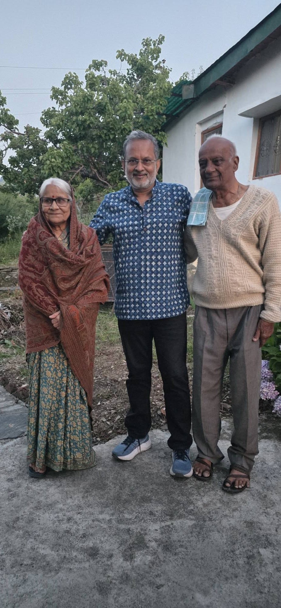 Three older adults stand together outdoors in a garden, posing for the photo in front of a house.